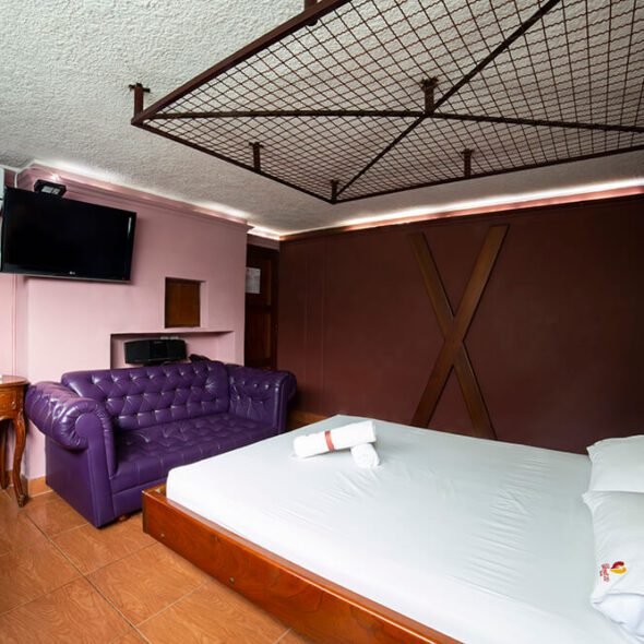 Un dormitorio de motel con una cama amplia y un sofá púrpura. Un televisor empotrado en la pared y un espejo redondo sobre un escritorio de madera complementan el espacio.