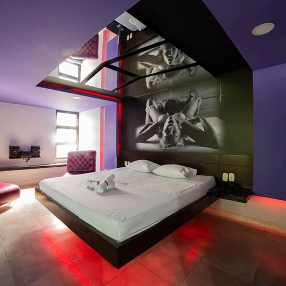 Un dormitorio de motel moderno iluminado por luces púrpuras, con una cama amplia y un impactante mural fotográfico en blanco y negro destacándose en la pared superior.