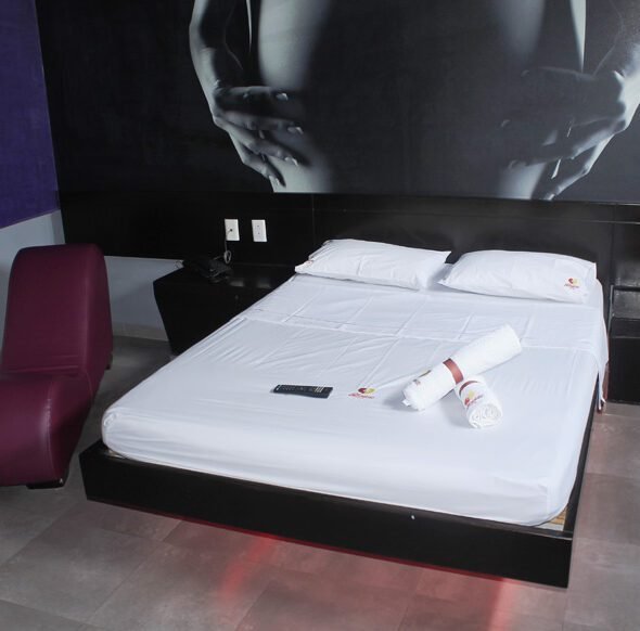 Elegante motel, ostenta cama grande con sábanas blancas, luces púrpuras dando un toque chic y una fotografía en blanco y negro.