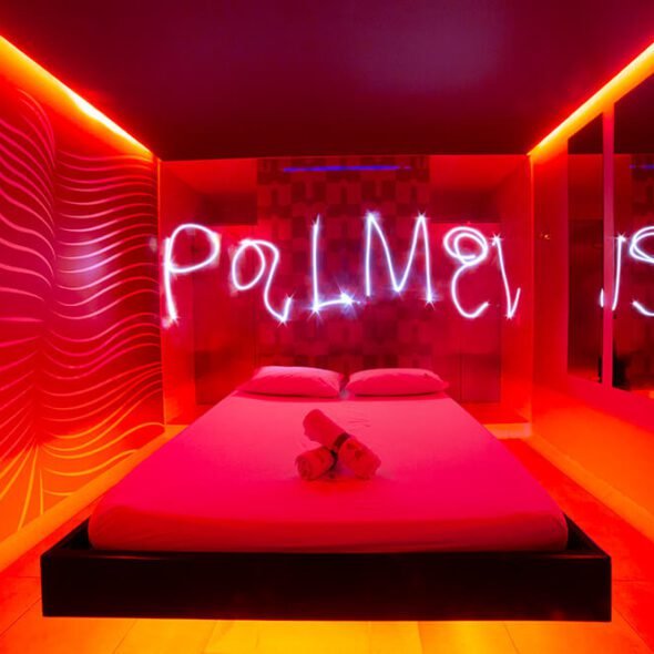 Habitación moderna de motel en Bucaramanga, bañada en luces de neón rojo con la palabra "poliamor" trazada en luz sobre la cama.