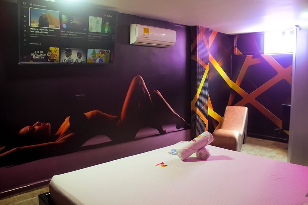 Un dormitorio moderno destaca arte en las paredes, ambiente de luz púrpura, TV colgada y un jacuzzi de motel.