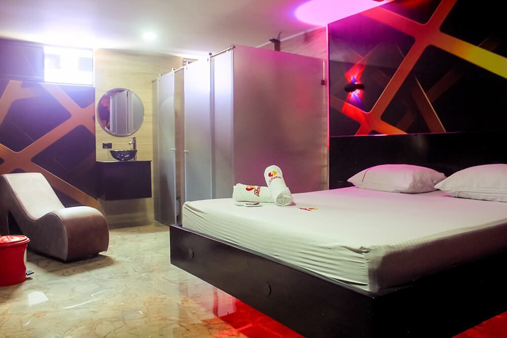 Moderno alojamiento de motel exhibiendo el lujo con una amplia cama, paredes artísticas, chaise lounge y un baño de cristal con jacuzzi.
