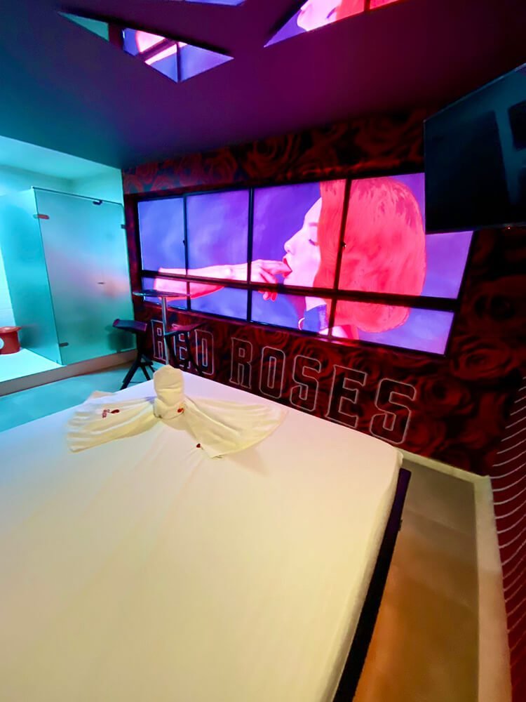Una vanguardista sala de karaoke en un motel con jacuzzi, bañada por luz roja, presenta un sofá blanco con cojín decorativo y una pantalla mostrando a un cantante.