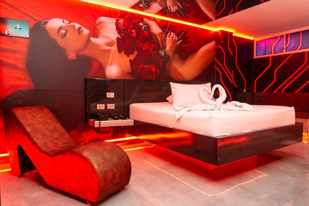 Un lujoso dormitorio con iluminación roja vibrante, amueblado al estilo moderno, exhibe un gran mural de una mujer entre flores y un jacuzzi del Motel.