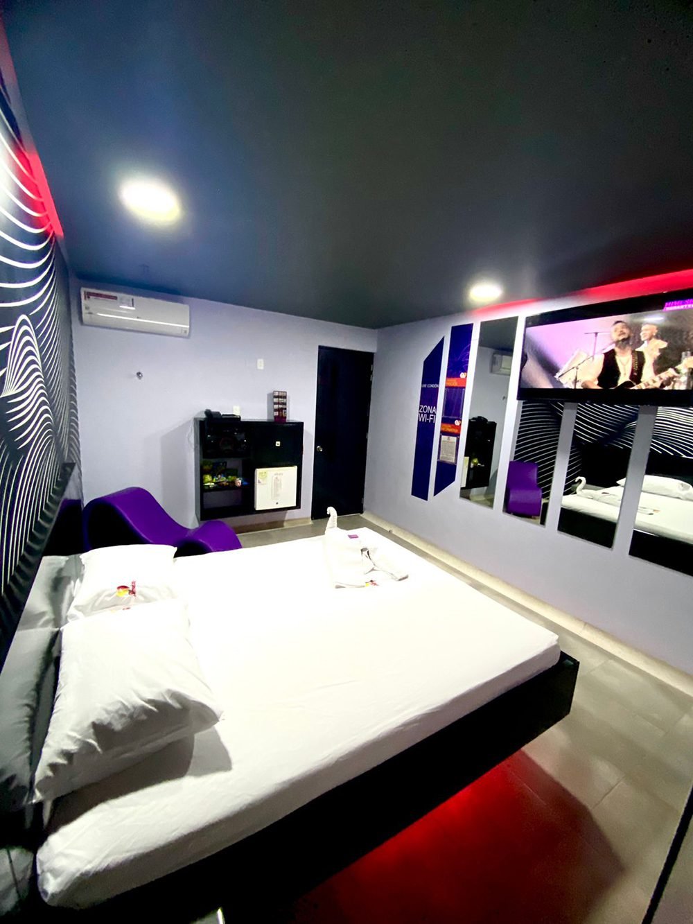 Dormitorio moderno, presenta una cama grande, acentos morados y un televisor fijo en la pared.