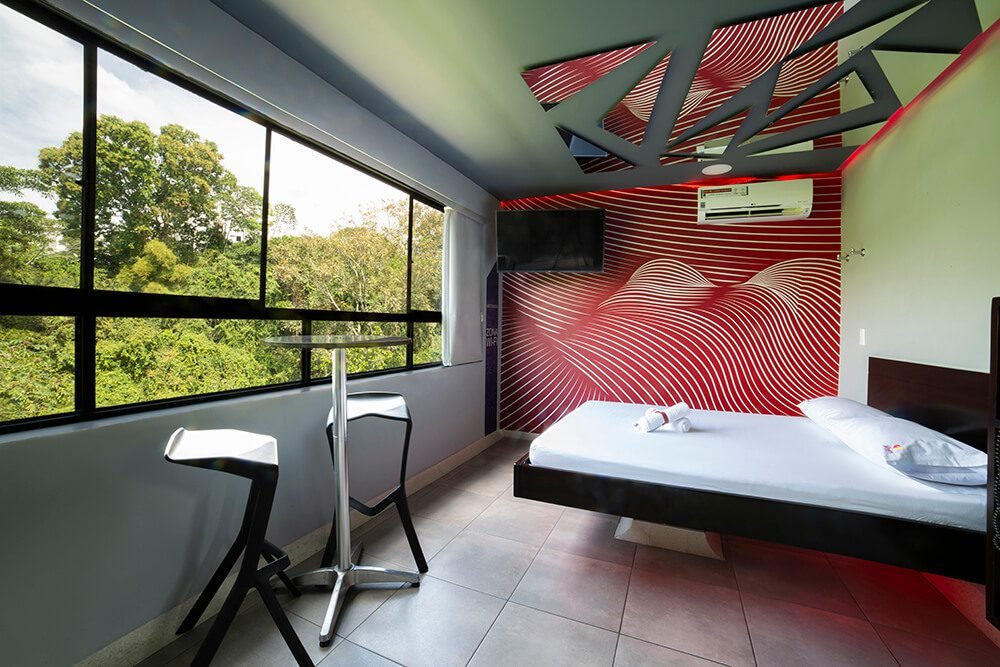 Un vívido motel presenta una audaz pared roja, ventana amplia con vista verde, mobiliario contemporáneo y un señorial jacuzzi.
